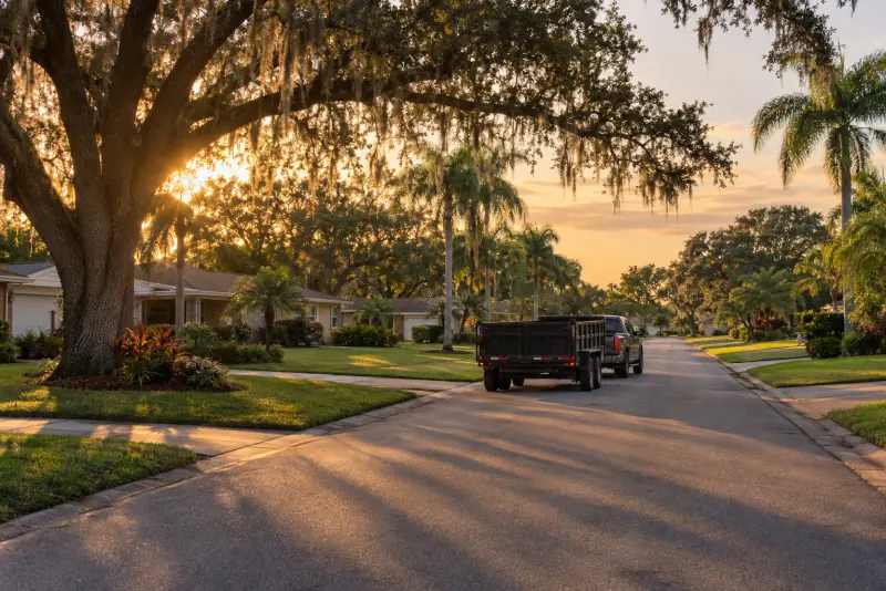 Pickup towing an empty dump trailer down a residential street lined with live oaks and palms at sunset